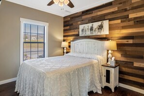 A bedroom with a bed, two bedside tables, a ceiling fan, a window with blinds, and a wooden wall panel with a painting. - The Fairview Hideaway (Fairview)