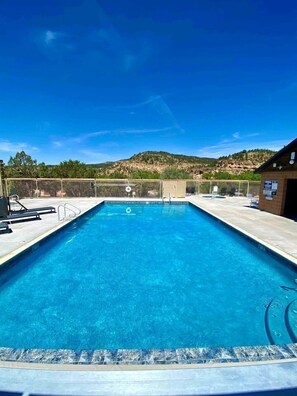 A rectangular swimming pool with clear blue water, surrounded by a fence, and a building with a brick exterior to the right. - New Modern Tiny House with Stargazing Windows in Orderville, Utah (Orderville)