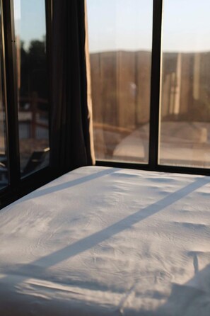 A bed with a white sheet, a window with a view of a fence, and curtains partially drawn. - New Modern Tiny House with Stargazing Windows in Orderville, Utah (Orderville)