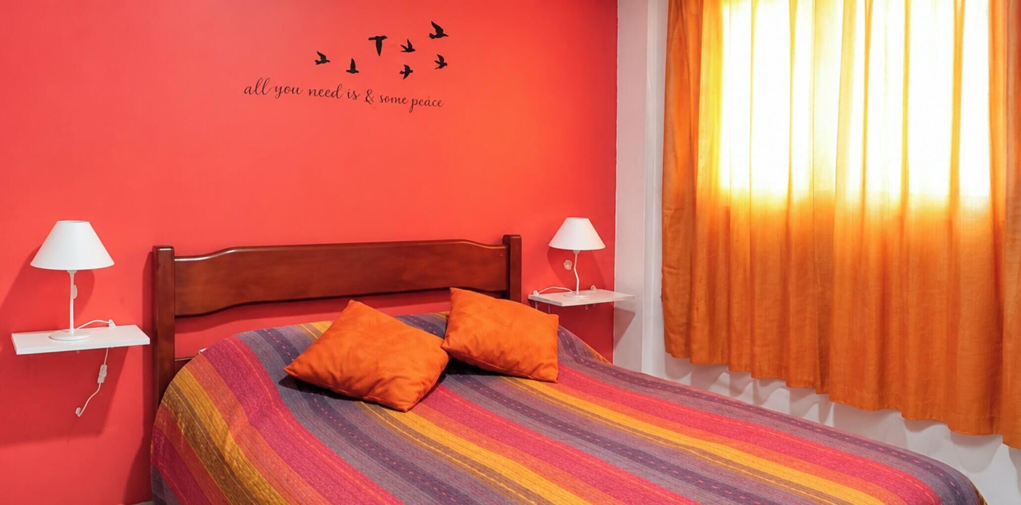 Een slaapkamer met een houten hoofdbord, twee nachtkastjes met lampen, een kleurrijke gestreepte spreidekbed, twee oranje kussens, en een muur met een silhouet van een vogel en tekst.