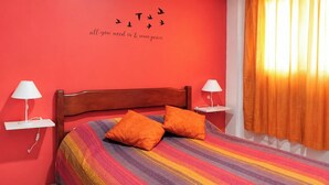 A bedroom with a wooden headboard, two bedside tables with lamps, a colorful striped bedspread, two orange pillows, and a wall with a bird silhouette and text. - Ap Balcan 500mts da Praia - Balneario Camboriu /SC (Balneário Camboriú)