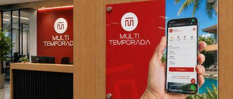 Zona de recepción de un hotel con un mostrador de madera, una pared roja con el logotipo del hotel y un cartel que muestra un teléfono inteligente con una interfaz de reservas.
