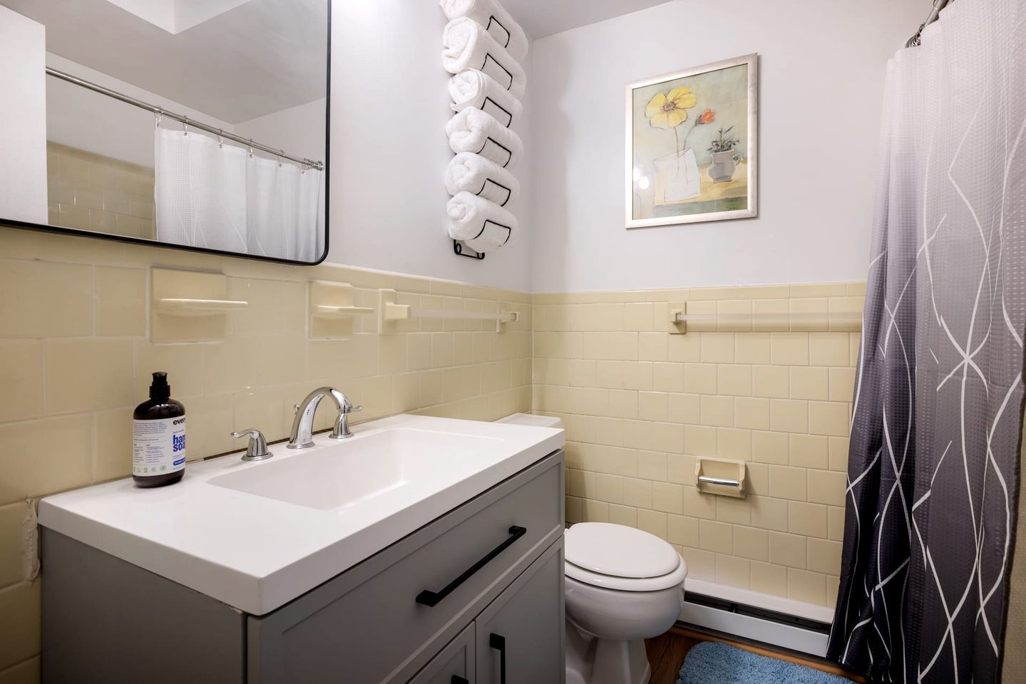 A bathroom with a white sink, two faucets, a toilet, a towel rack with folded towels, a shower curtain, and a framed picture on the wall.