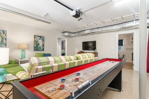 A room with a foosball table, a sofa, a television, and a projector mounted on the ceiling. - Salt & Serenity | Sleeps 14·Game Room·Walk to Beach (Pine Knoll Shores)