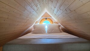 A narrow, wooden-paneled attic bedroom with a sloped ceiling, a bed with white linens, and a circular skylight.