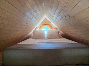 A narrow, wooden-paneled attic bedroom with a sloped ceiling, a bed with white linens, and a circular skylight. - Ailéa – Tiny House with a Private Hot Tub in Provence (Beaumes-de-Venise)