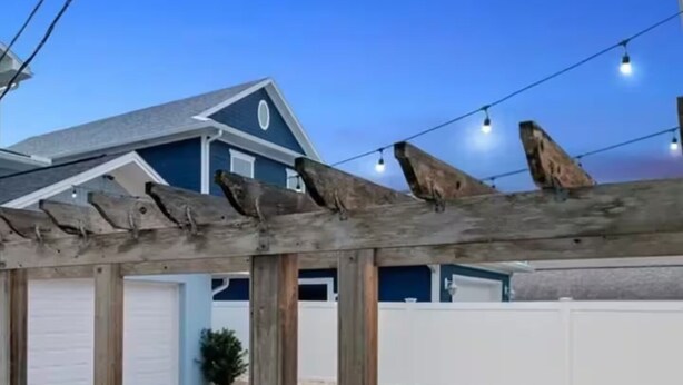 Eine Pergola aus Holz, gestützt von Holzsäulen, mit einem Kiesweg und einem blauen Gebäude im Hintergrund.