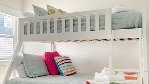 Ein Stockbett mit weißem Gestell, grauer Bettwäsche und einem Fenster mit Jalousien.