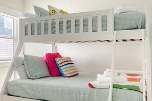 Ein Stockbett mit weißem Gestell, grauer Bettwäsche und einem Fenster mit Jalousien.