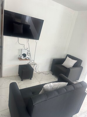 A living room with a flat-screen TV mounted on the wall, two black armchairs, a small ottoman, and a white tiled floor.