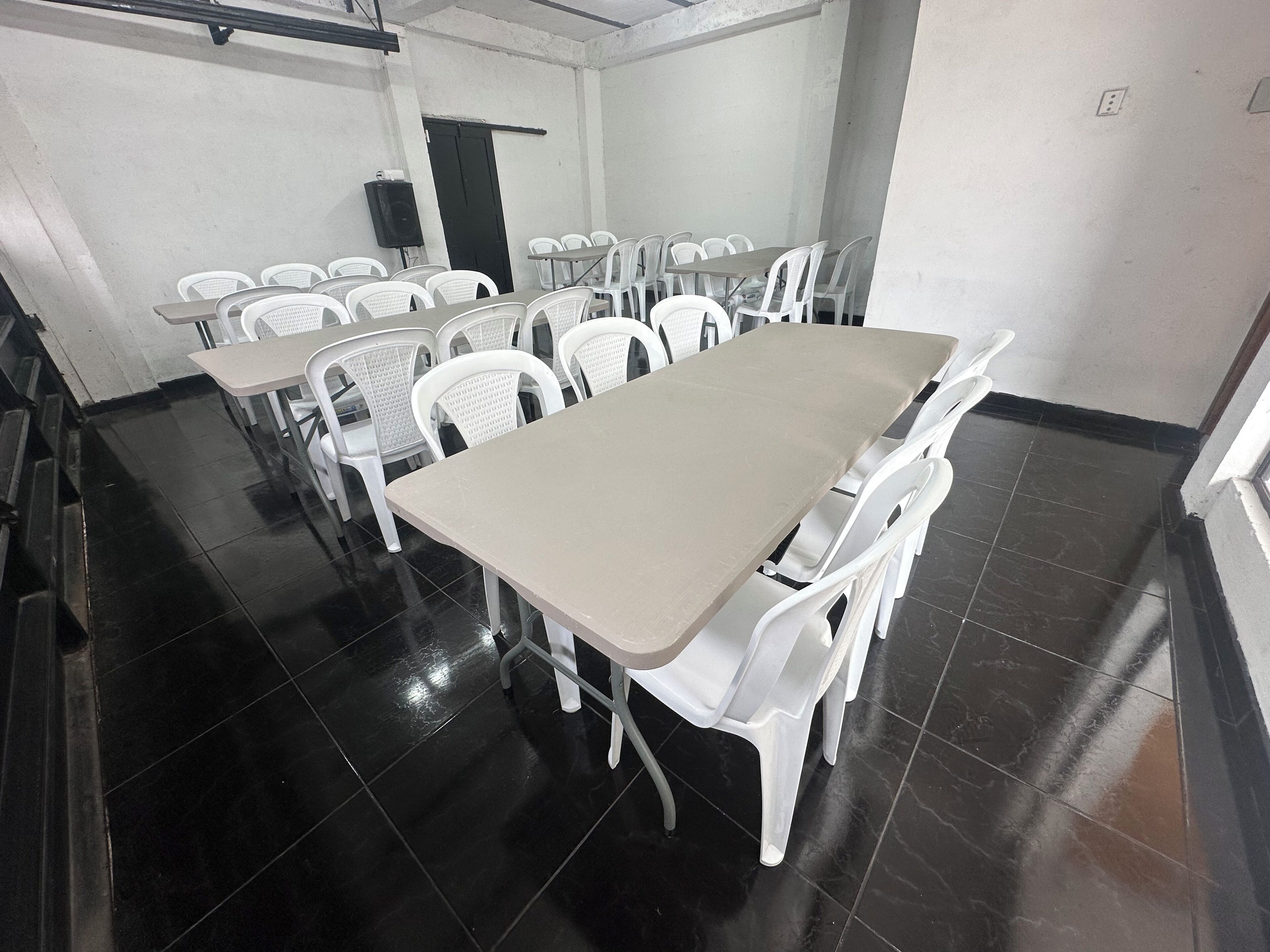 Ein Konferenzraum mit einem langen Tisch, weißen Stühlen, einer schwarzen Tür und einem Redner in der Ecke.