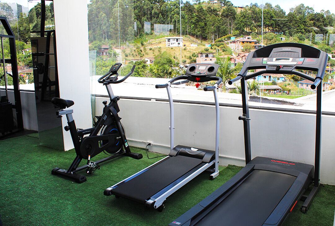 Een sportschool met een loopband, elliptische trainer en hometrainer, met uitzicht op een woongebied.