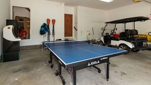 A ping pong table in a room with a golf cart, a table tennis table, and a door. - The Grand Carolina | Golf Cart ·Hot Tub ·4BR ·PKS (Pine Knoll Shores)
