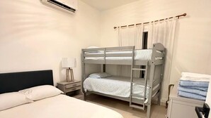 A hotel room with a bunk bed, a single bed, an air conditioning unit, a bedside lamp, and a cabinet.