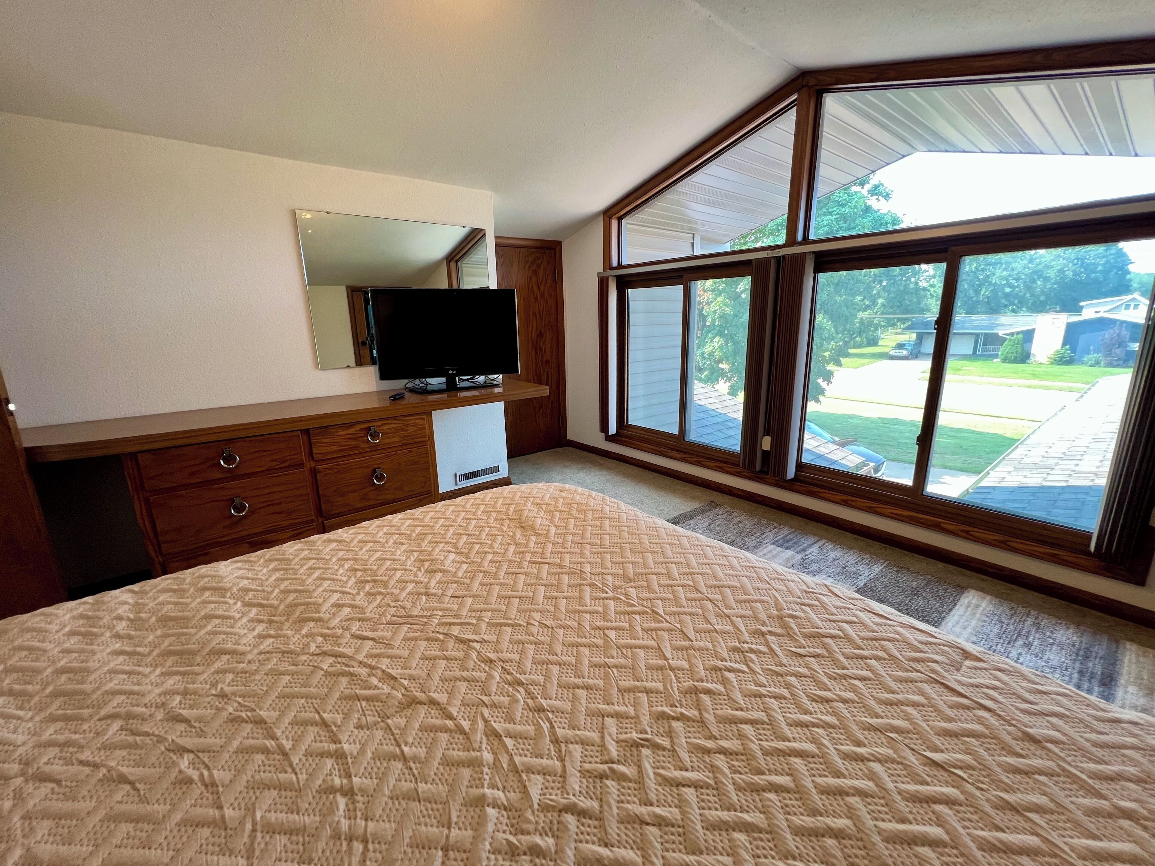 Een slaapkamer met een groot bed, een dressoir, een televisie en een raam met uitzicht op een woonwijk.