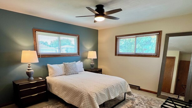 A bedroom with a bed, two bedside tables with lamps, a ceiling fan, and two windows with blinds. - Spacious Independence Home - Conveniently Located (Independence)