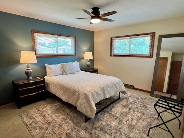 A bedroom with a bed, two bedside tables with lamps, a ceiling fan, and two windows with blinds.