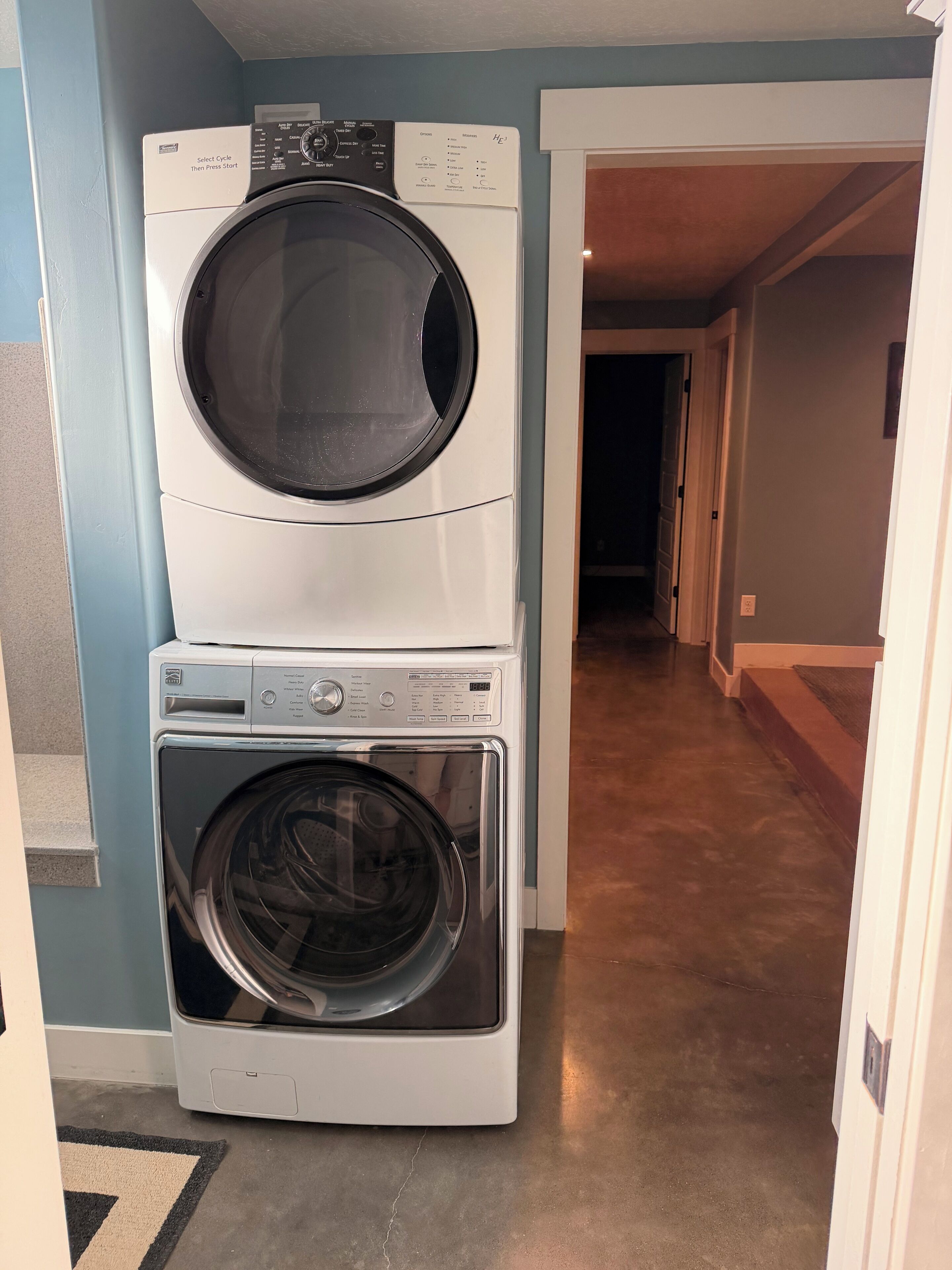 A front-loading washer and dryer stacked in a laundry room with a hallway leading to another room.
