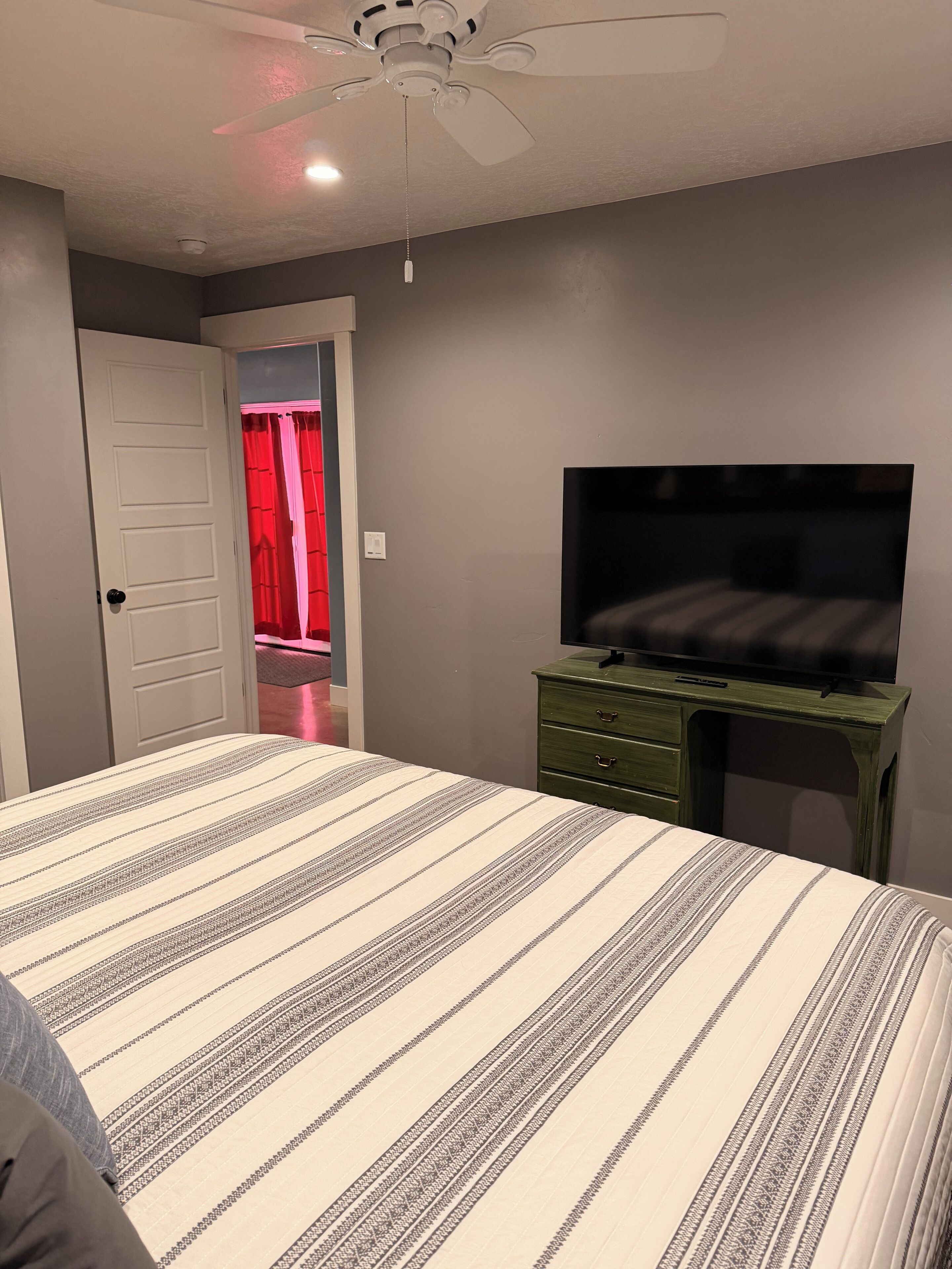 A bedroom with a bed, a desk, a television, and a ceiling fan.