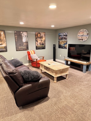 A living room with a brown leather couch, a red armchair, a wooden coffee table, a TV, and movie posters on the wall.