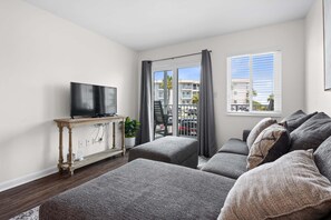 Un salon moderne avec un téléviseur à écran plat posé sur une console en bois, des sièges recouverts d’un tissu gris, une plante en pot et une vue sur un balcon donnant sur des voitures et des immeubles.