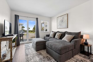 Ein modernes Wohnzimmer mit einem grauen Sofa, einem Fernseher auf einem Holzboden, einem kleinen Tisch und Blick ins Freie durch eine Glastür.