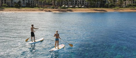 Two people paddleboarding on a clear blue water body with a resort in the background.
