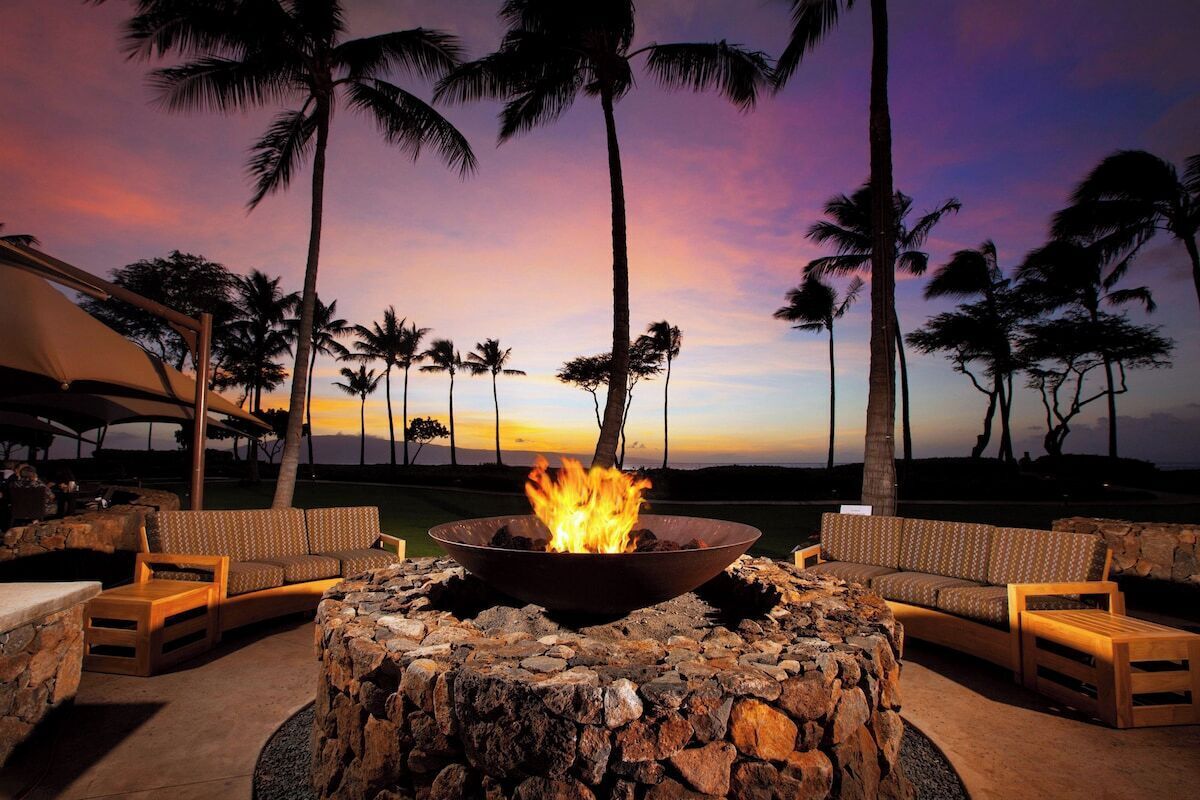 A fire pit surrounded by stone seating, palm trees, and a sunset view.