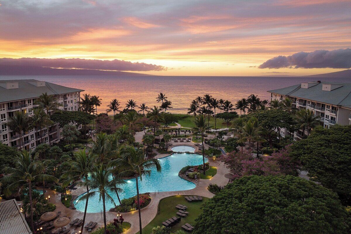 A resort with a swimming pool, palm trees, and a view of the ocean at sunset.