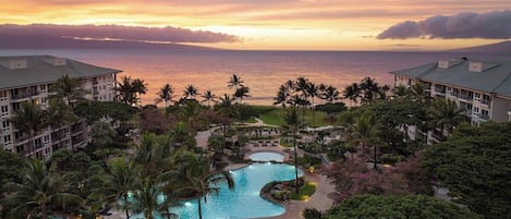 A resort with a swimming pool, palm trees, and a view of the ocean at sunset.