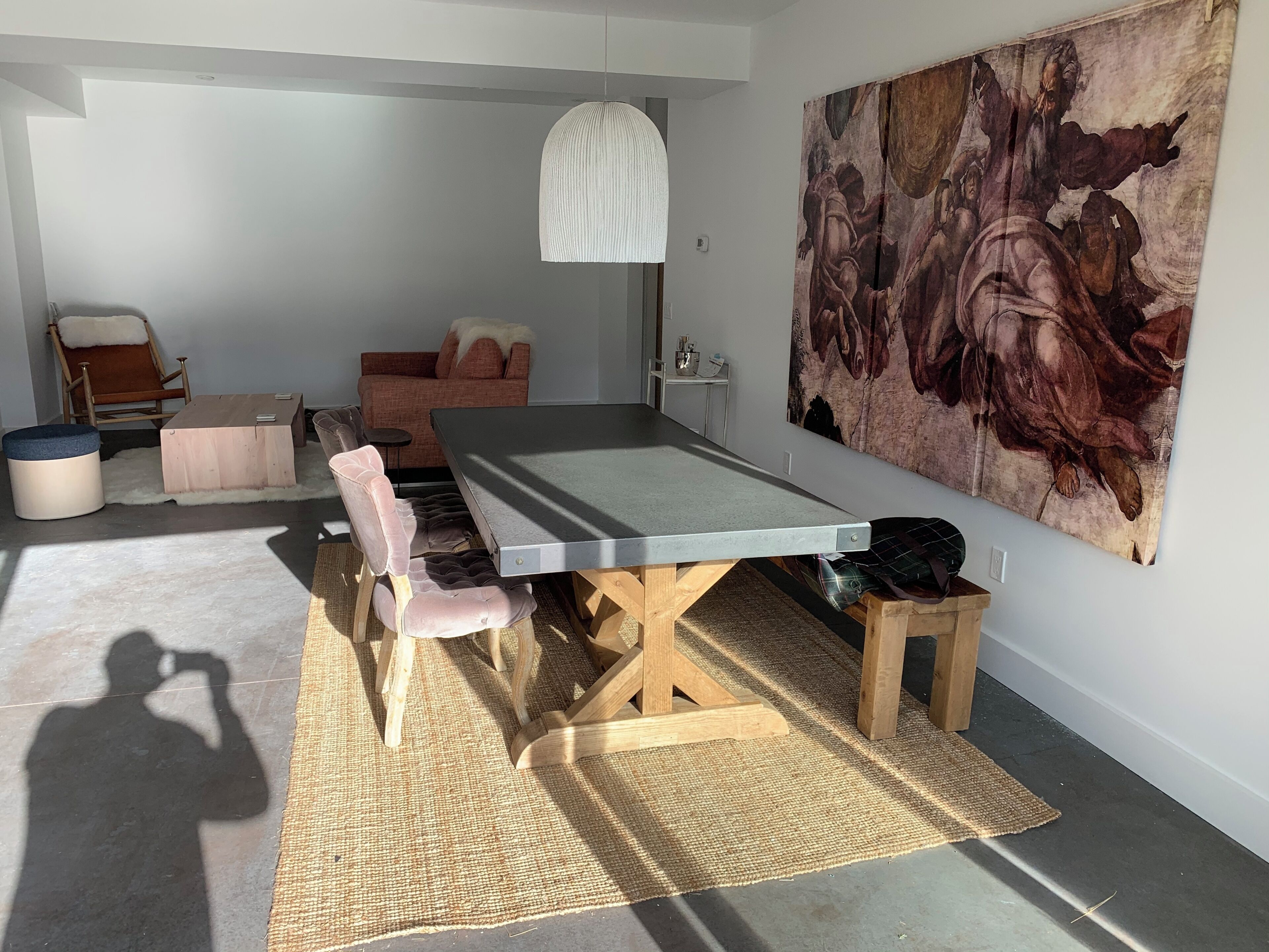 Une pièce avec une longue table, des chaises rembourrées, un luminaire suspendu et un grand tableau accroché au mur.