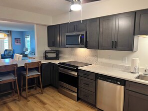 A modern kitchen with dark cabinets, a microwave, a toaster, a dishwasher, and a dining area with a table and chairs. - Sea Oats A102 by ALBVR - Lagoon Front Updgraded Condo (Gulf Shores)