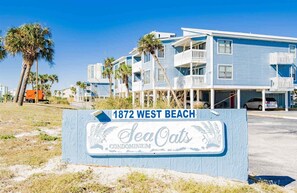 Un complejo de condominios frente a la playa llamado 'Sea Oats', con varias edificaciones, un cartel que indica '1872 West Beach' y un cielo azul despejado.