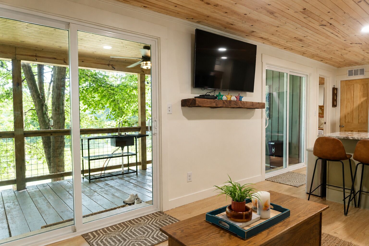 A modern living room with a large TV, wooden ceiling, sliding glass doors leading to a patio, and a coffee table with decorative items.