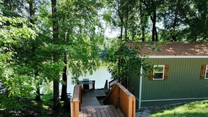 A wooden deck leading to a cabin with a dock extending into a lake, surrounded by dense greenery.