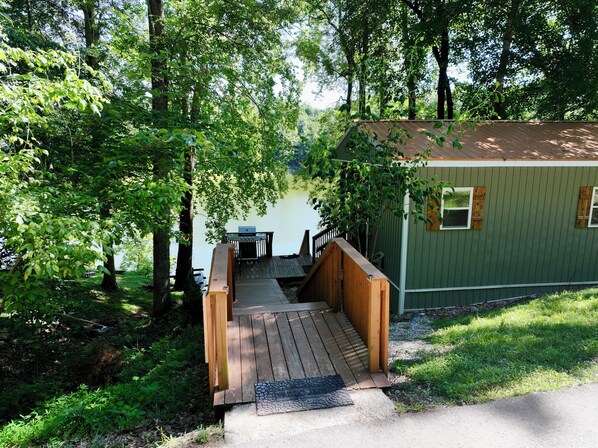 A wooden deck leading to a cabin with a dock extending into a lake, surrounded by dense greenery.