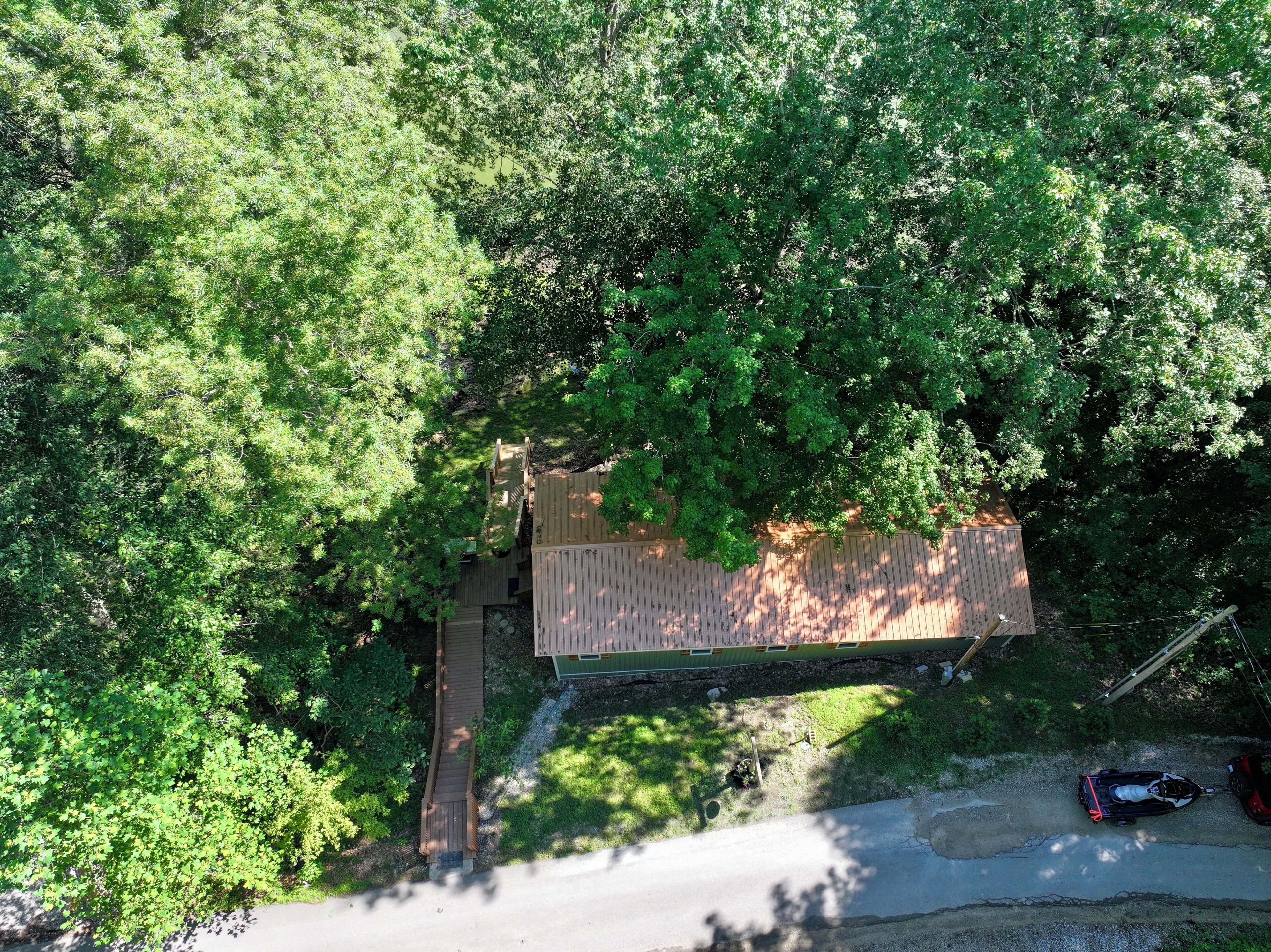 A small cabin surrounded by dense trees, with a wooden deck and a paved driveway.