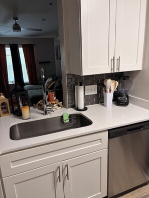 Eine Küche mit Spülbecken aus Edelstahl, einem Mixer und einer Kaffeemaschine. Über der Arbeitsplatte befinden sich Schränke und über dem Raum ein Deckenventilator.
