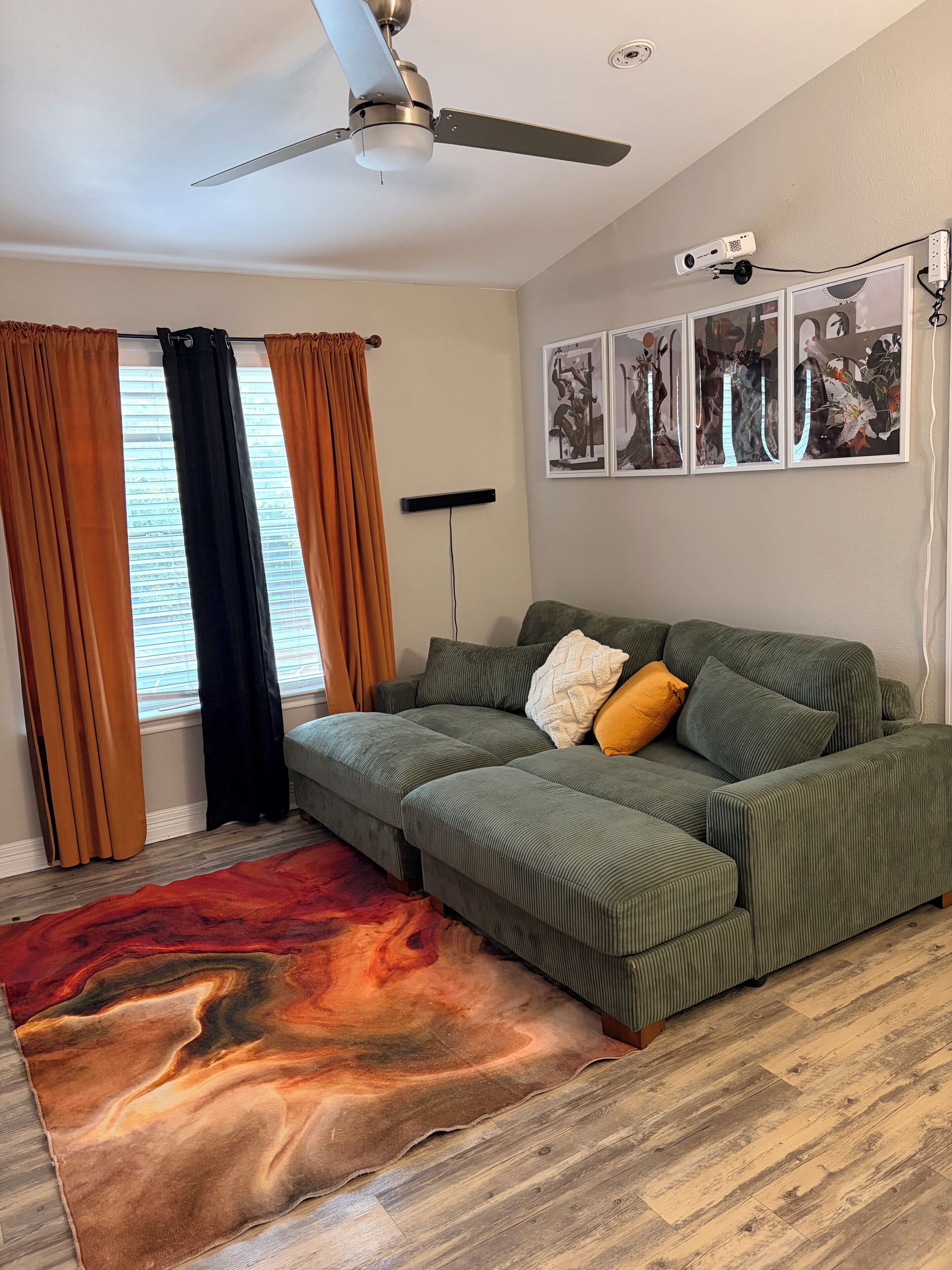 Una sala de estar con un sofá verde, un tapete rojo y naranja, un ventilador de techo y un proyector en la pared.