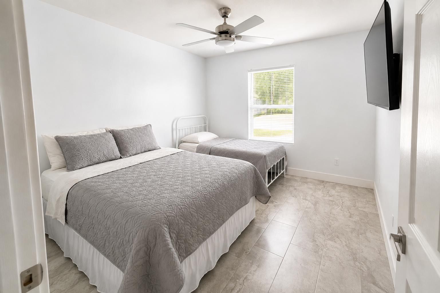 Une chambre avec deux lits, un ventilateur de plafond, une fenêtre avec des stores et un téléviseur fixé au mur.