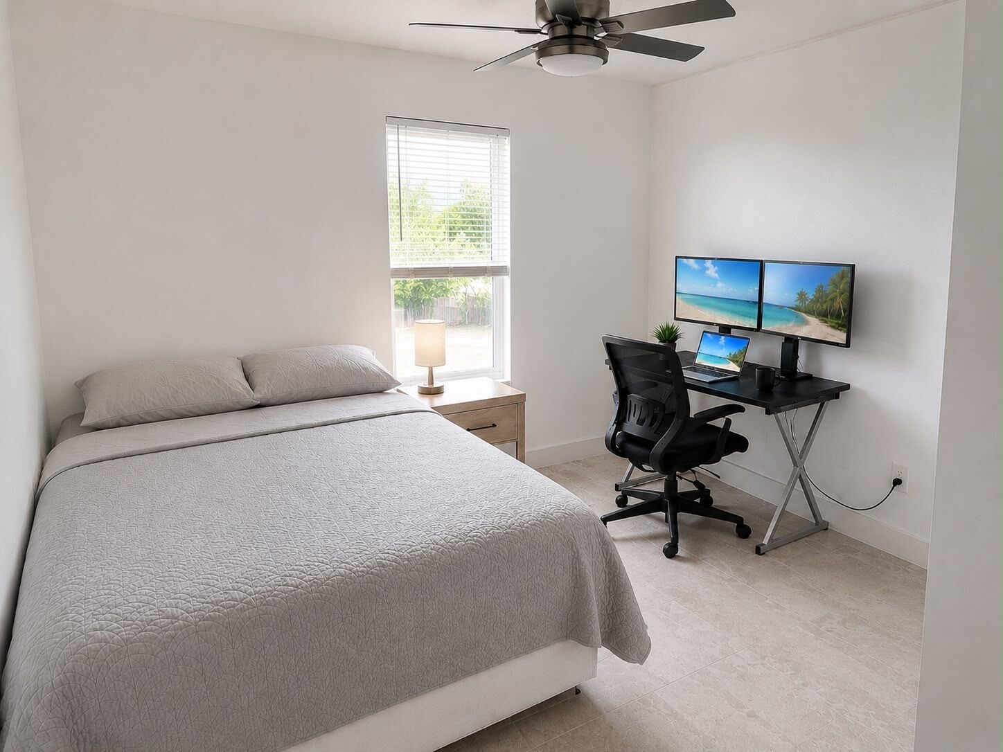 Quarto com cama, escrivaninha com computador e dois monitores, ventilador de teto e janela com persianas.