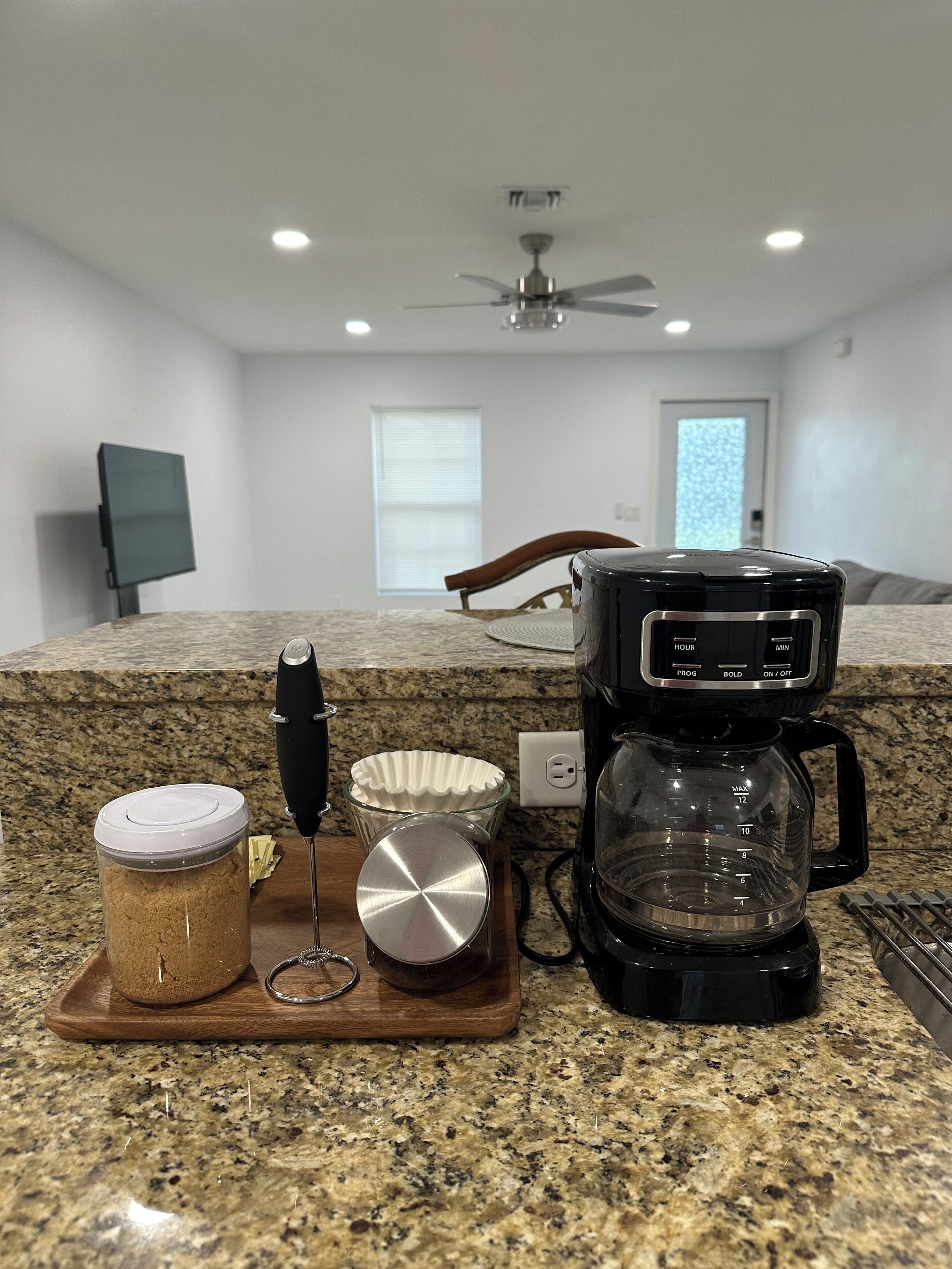 A kitchen counter with a coffee maker, a jar of sugar, a metal whisk, and a glass measuring cup.