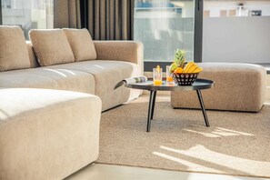 Ein modernes Wohnzimmer mit einem beigefarbenen Sofa, einem runden Couchtisch mit einer Schale voller Obst und Getränke, und einem großen Fenster, das für natürliches Licht sorgt.
