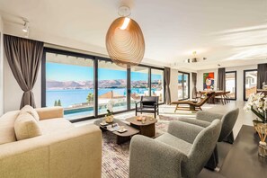 Ein geräumiges Wohnzimmer mit großem Fenster, das Blick auf ein Gewässer und Berge bietet, einer modernen Kronleuchter und einem Essbereich mit Aussicht.