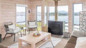 Interior acogedor de una cabaña con chimenea, dos sillones, un sofá y ventanas amplias con vistas al exterior.