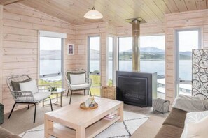 A cozy cabin interior with a fireplace, two chairs, a sofa, and large windows offering a view of the outdoors. - Aotea Harbour Beach House (Kawhia)