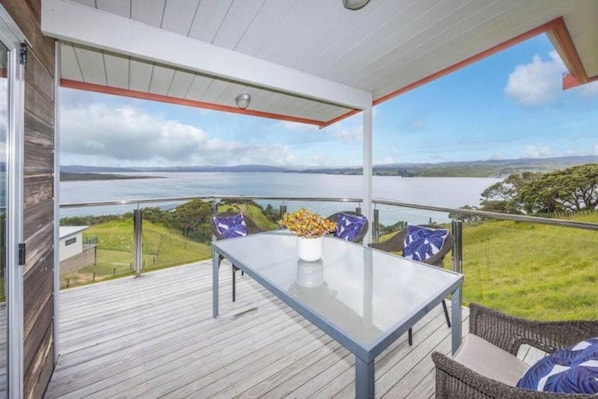 A balcony with a glass table and chairs, overlooking a body of water and greenery.