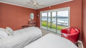 Un dormitorio con dos camas, una silla roja, un ventilador de techo y un ventanal con vista a un edificio y al agua.