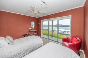 A bedroom with two beds, a red chair, a ceiling fan, and a large window with a view of a building and water. - Aotea Harbour Beach House (Kawhia)