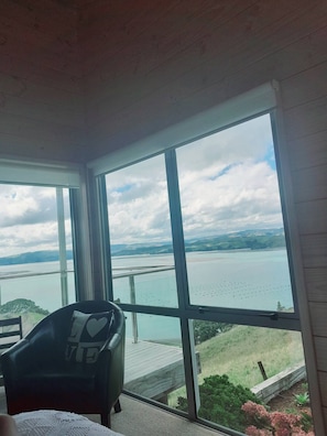 A room with a large window offering a view of a lake and distant hills. - Aotea Harbour Beach House (Kawhia)